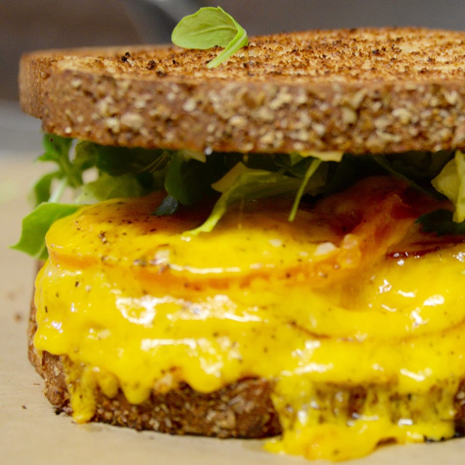 Close-up of a grilled cheese sandwich with melted yellow cheese, tomatoes, and greens on toasted bread.