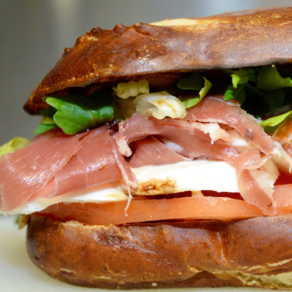 Close-up of a Sutter Street Cafe sandwich with prosciutto, mozzarella, tomato, and lettuce on a pretzel bun.